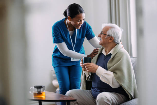 Caregiver helping an older adult hold a warm mug at a sunlit kitchen table